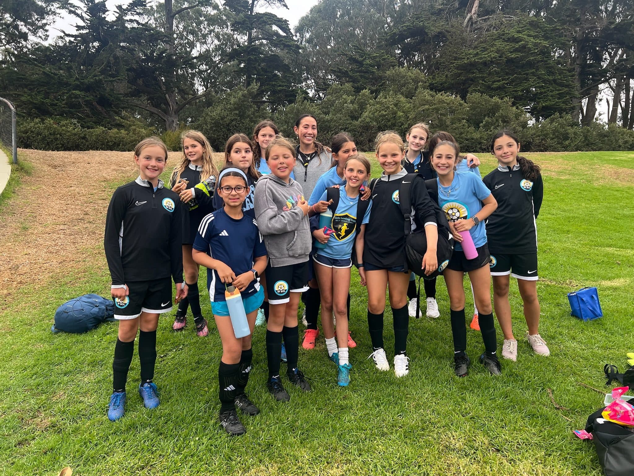 About fourteen girls in a mix of black GLG kits, warmups and light blue tees pose smiling on grass beneath park trees, several holding water bottles