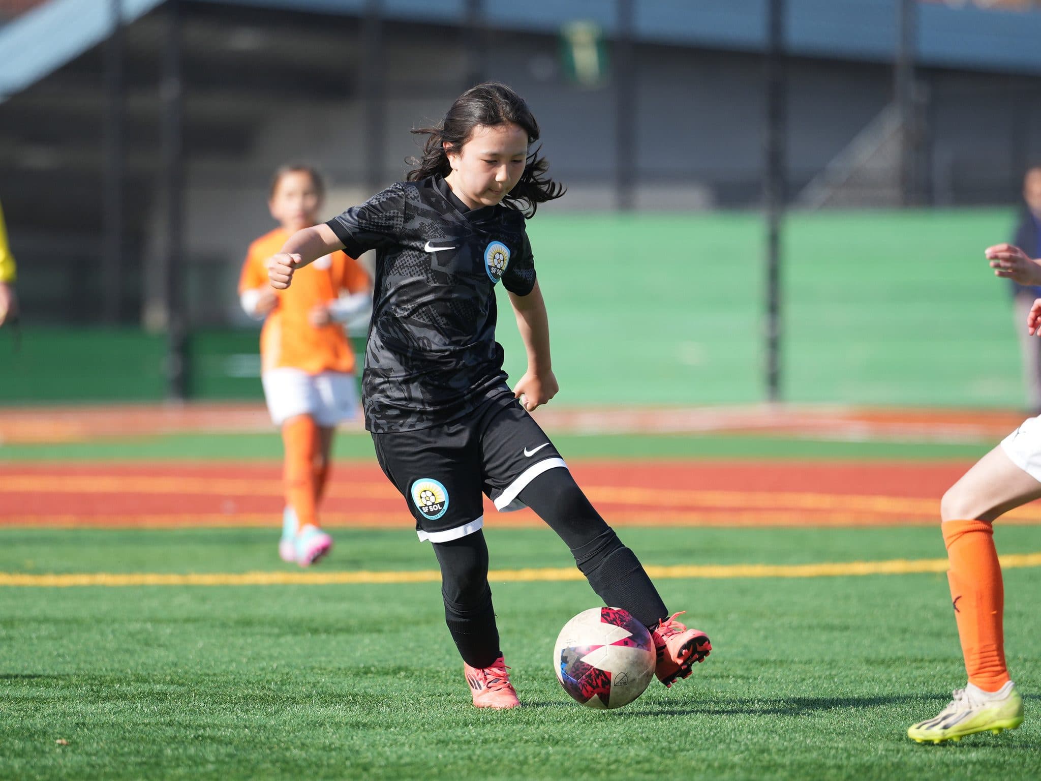 A GLG player in a black kit controls the ball mid-dribble while two opponents in orange close in on a red-and-green turf field