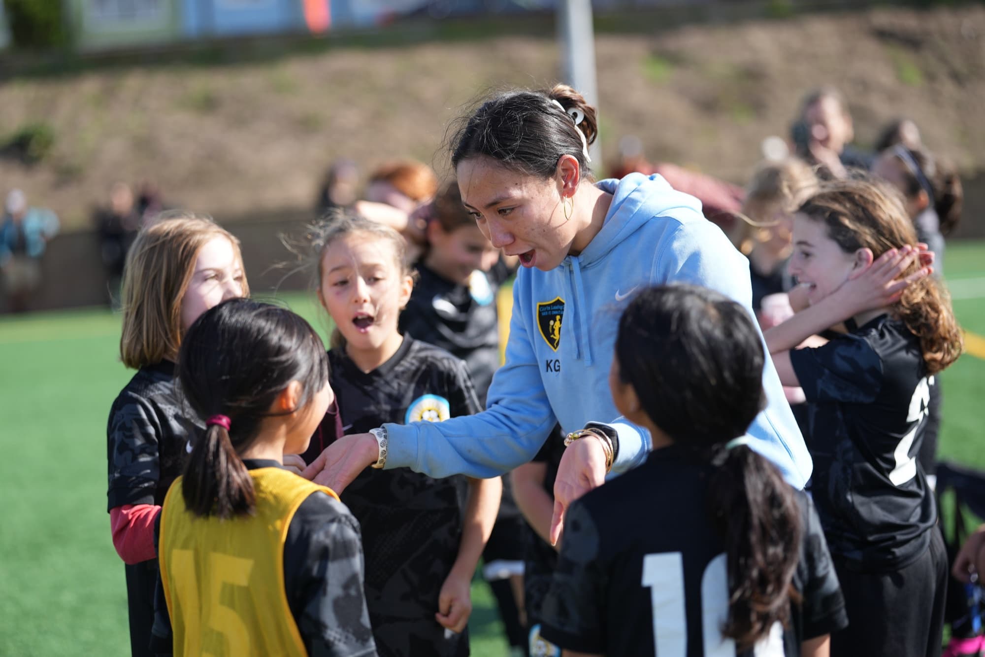 A coach in a light blue SF SOL hoodie leans into a circle of players, talking animatedly as they listen wide-eyed