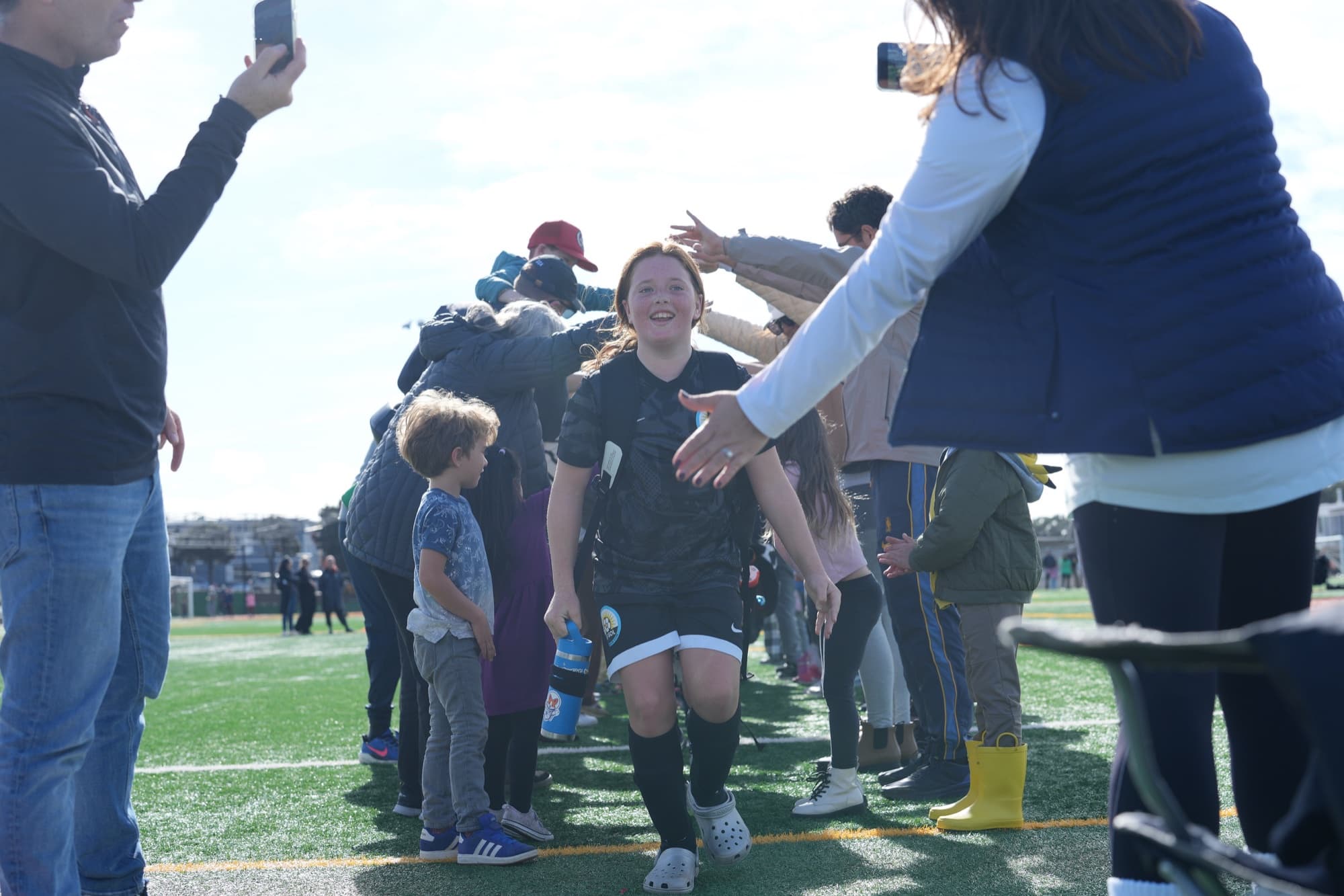 A smiling player in a black kit jogs through a tunnel of parents reaching out for high fives, phones raised to film her