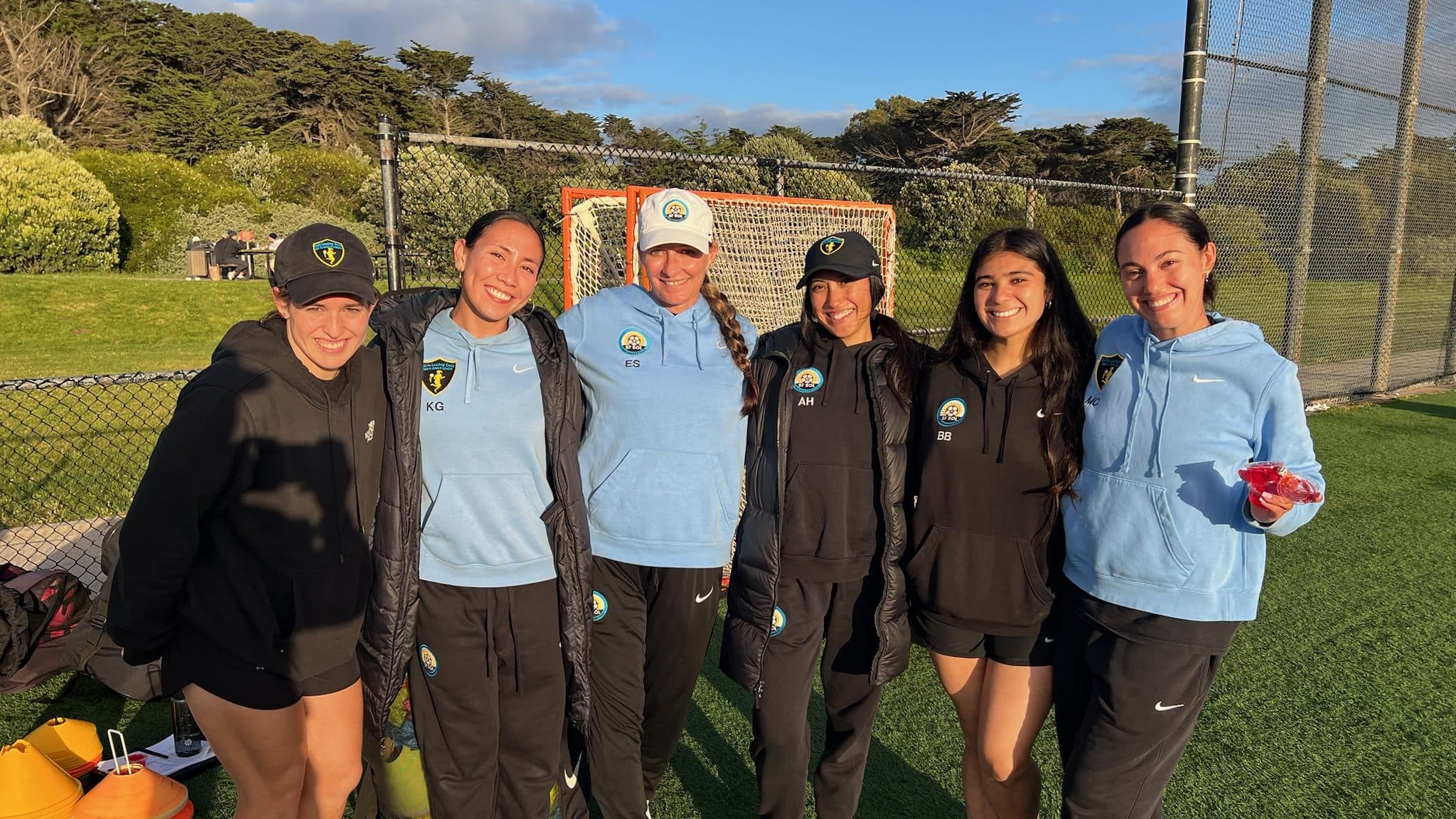 Six SF SOL coaches in matching tracksuits and hoodies, smiling together on the field in golden-hour light
