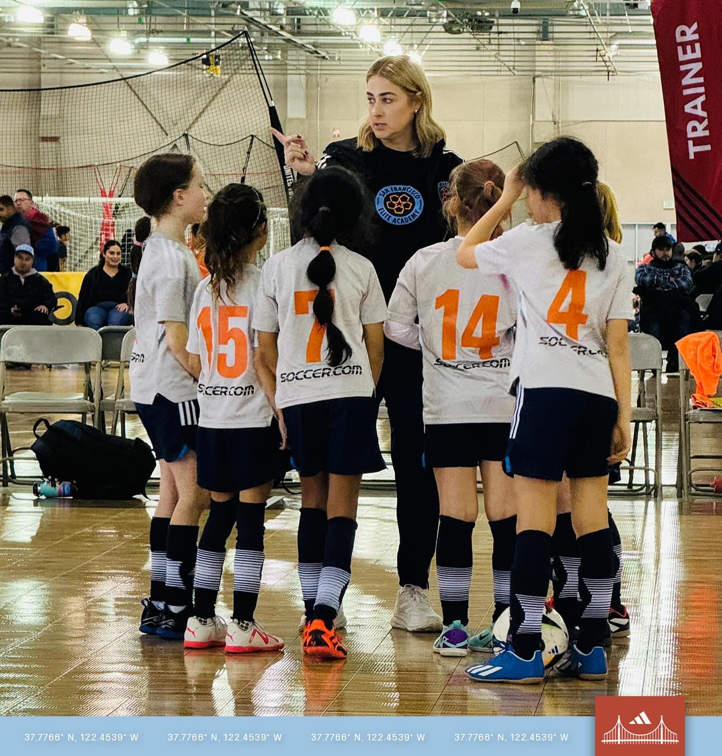 Loni crouches with a hand raised, giving focused instructions to her numbered players huddled around her on the sideline of an indoor court