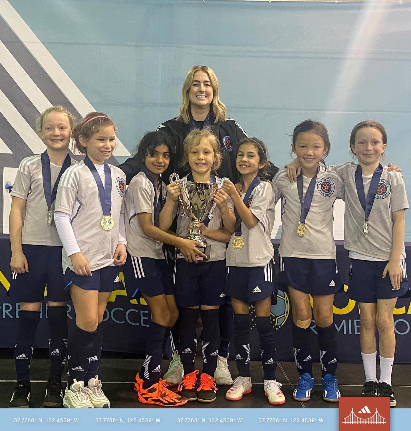 Loni's team lines up smiling in matching white and navy kits with medals around their necks, Loni standing behind holding a trophy overhead with a player, after an indoor tournament win
