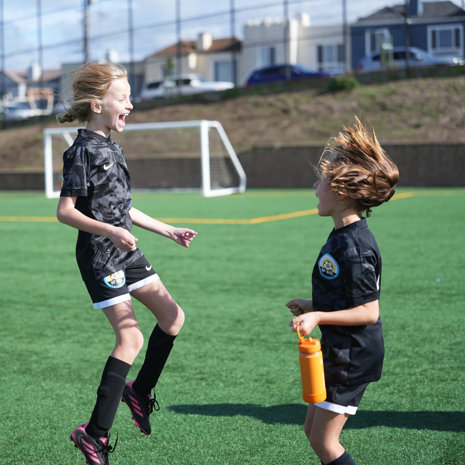Two GLG players in black kits leap and laugh mid-air in front of an open goal, one clutching an orange water bottle, SF houses on the hillside behind