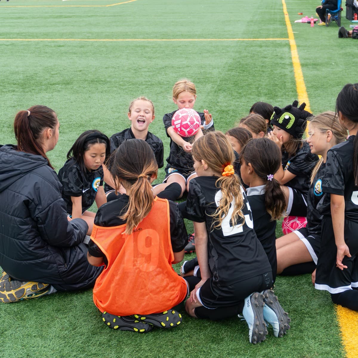 A coach kneels on the turf with her team in black kits gathered around her on the sideline, one player laughing mid-sentence, another holding up a pink ball