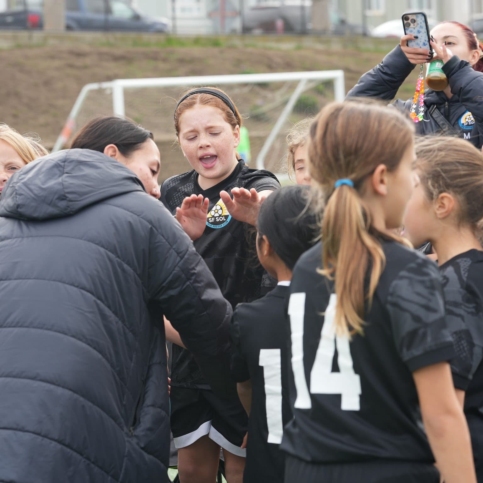 A coach in a black jacket huddles closely with excited players in black number jerseys, one player mid-cheer, while a parent captures the moment on a phone behind them