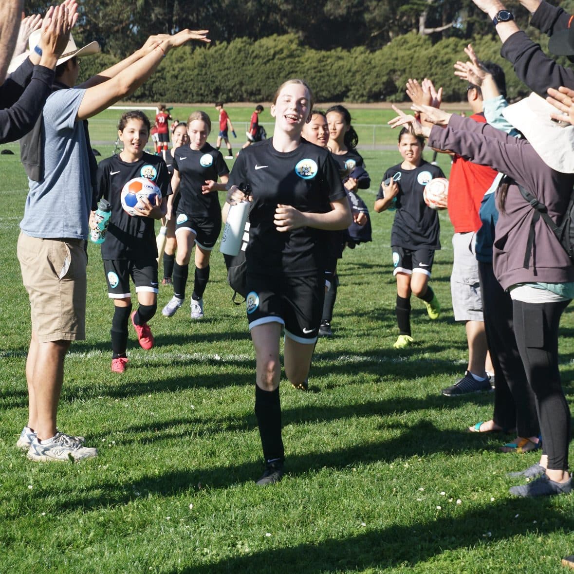 Vertical frame of the whole team in black kits running one by one through a tunnel of clapping parents on a grass field, the lead player smiling wide