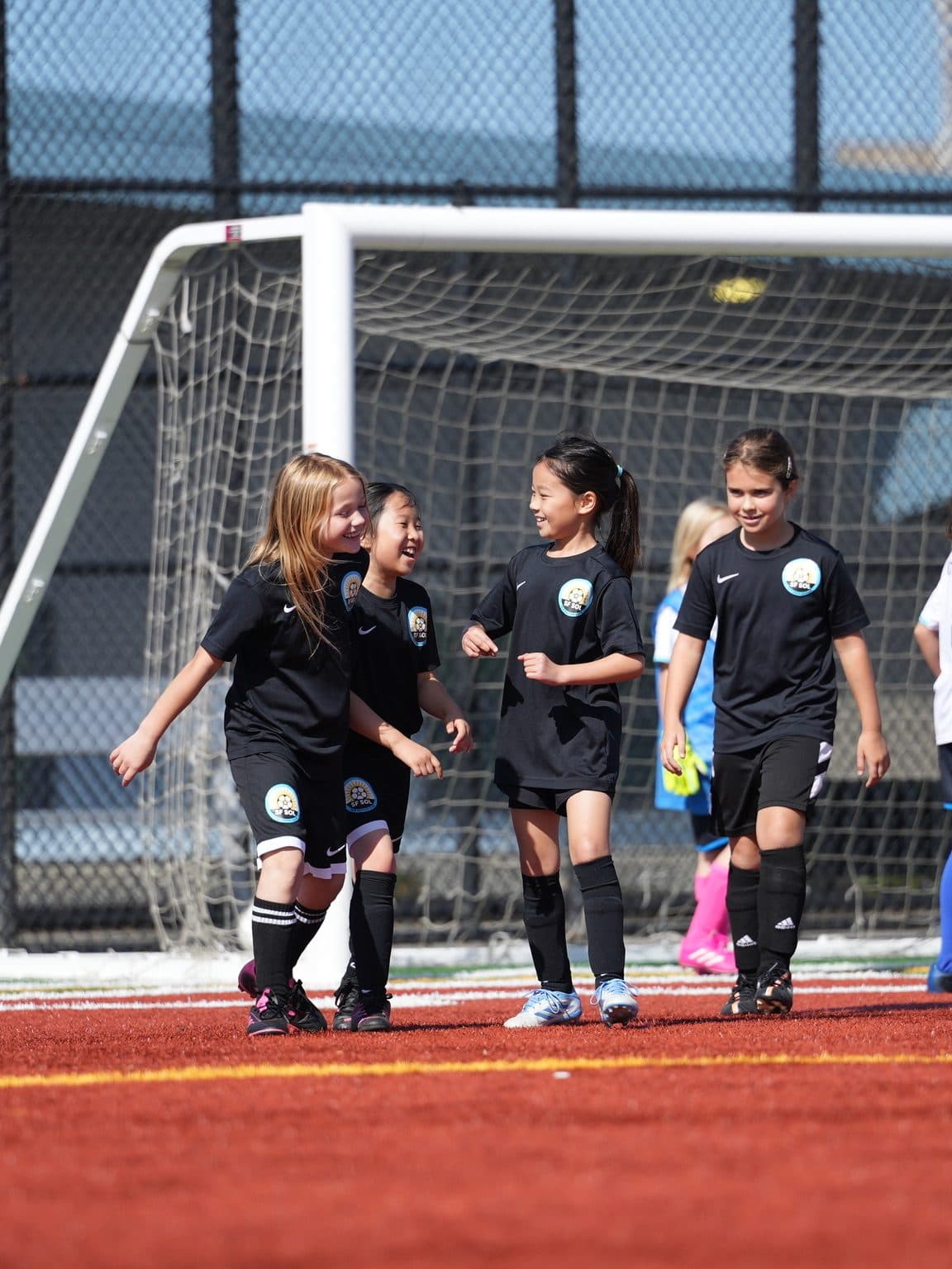 Four girls in black kits walk shoulder-to-shoulder off the pitch smiling, with opponents in white and a goal frame behind them