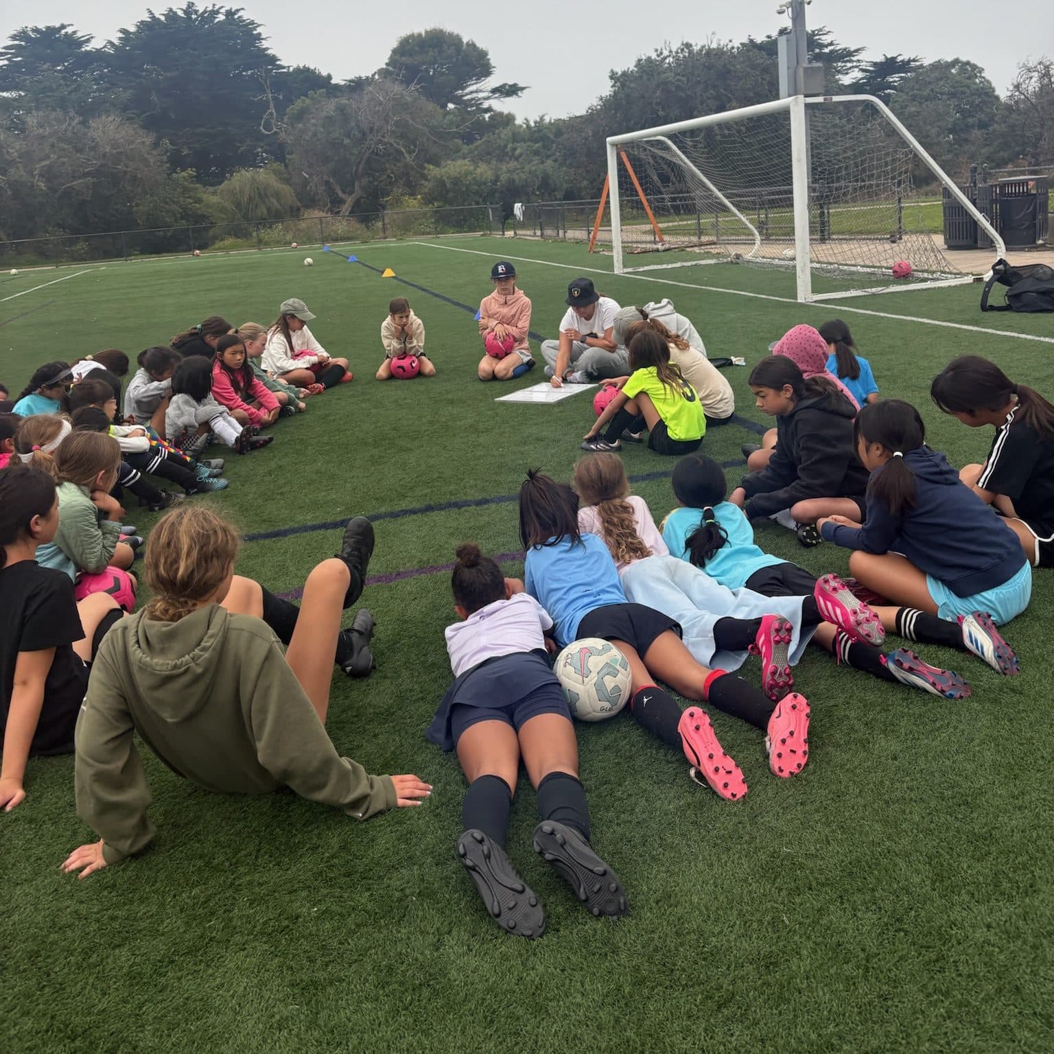A big group of campers in casual and colorful clothes sits and sprawls in a circle on a foggy turf field while coaches in caps lead the discussion near the goal