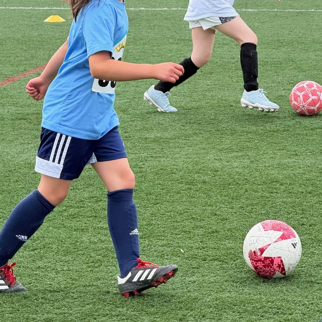 Vertical practice shot of a young player in a light blue rec tee mid-dribble, ponytail flying, with a second player in a white pinnie dribbling behind and more kids across the turf