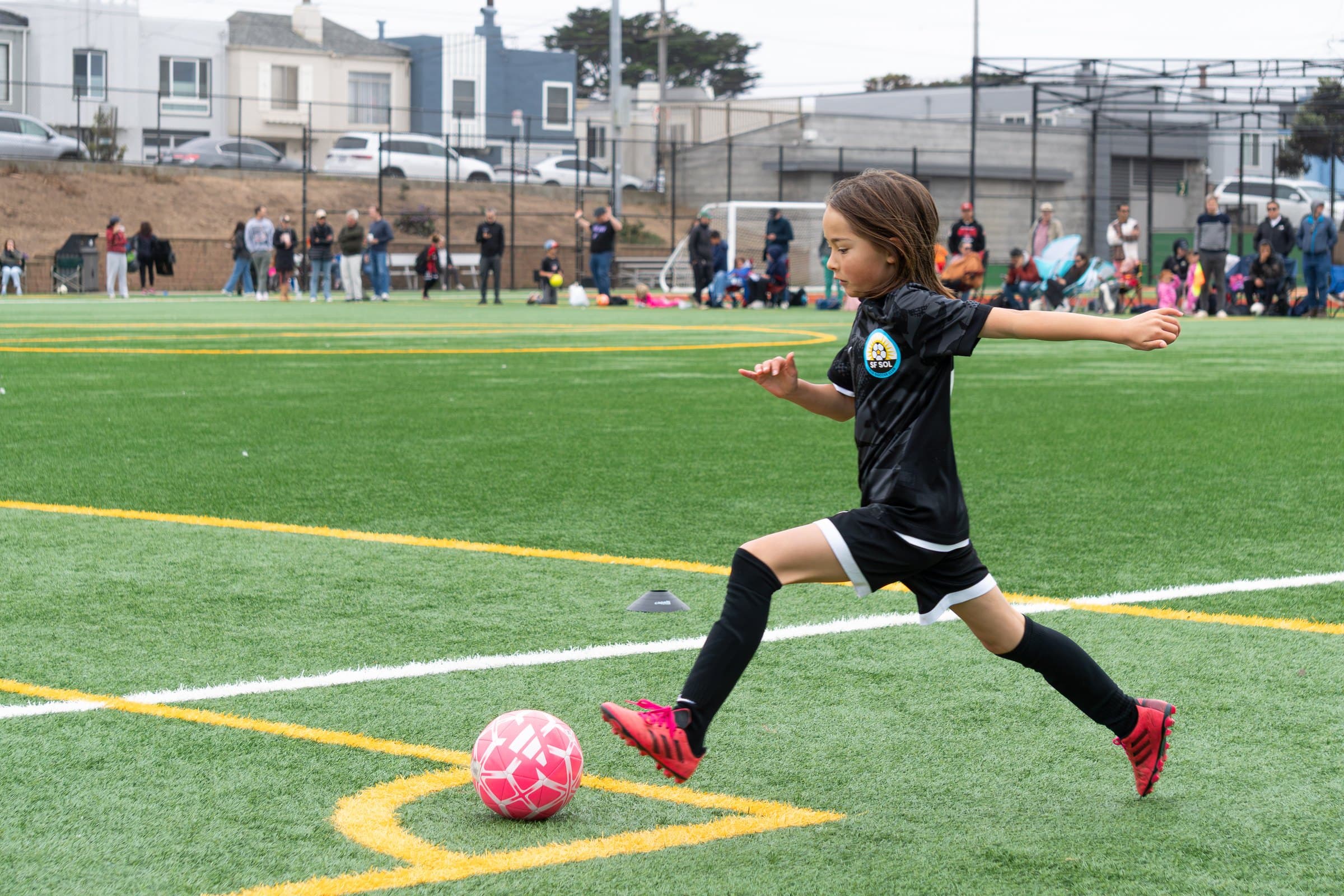 A player in a black kit stretches into a corner kick with a pink ball, houses and spectators along the far side of the field