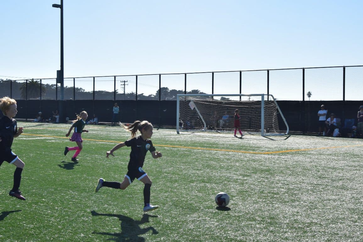 A player in a black kit sprints hard toward a loose ball in front of the goal on a sunlit turf field, hair flying, with teammates and opponents trailing the play