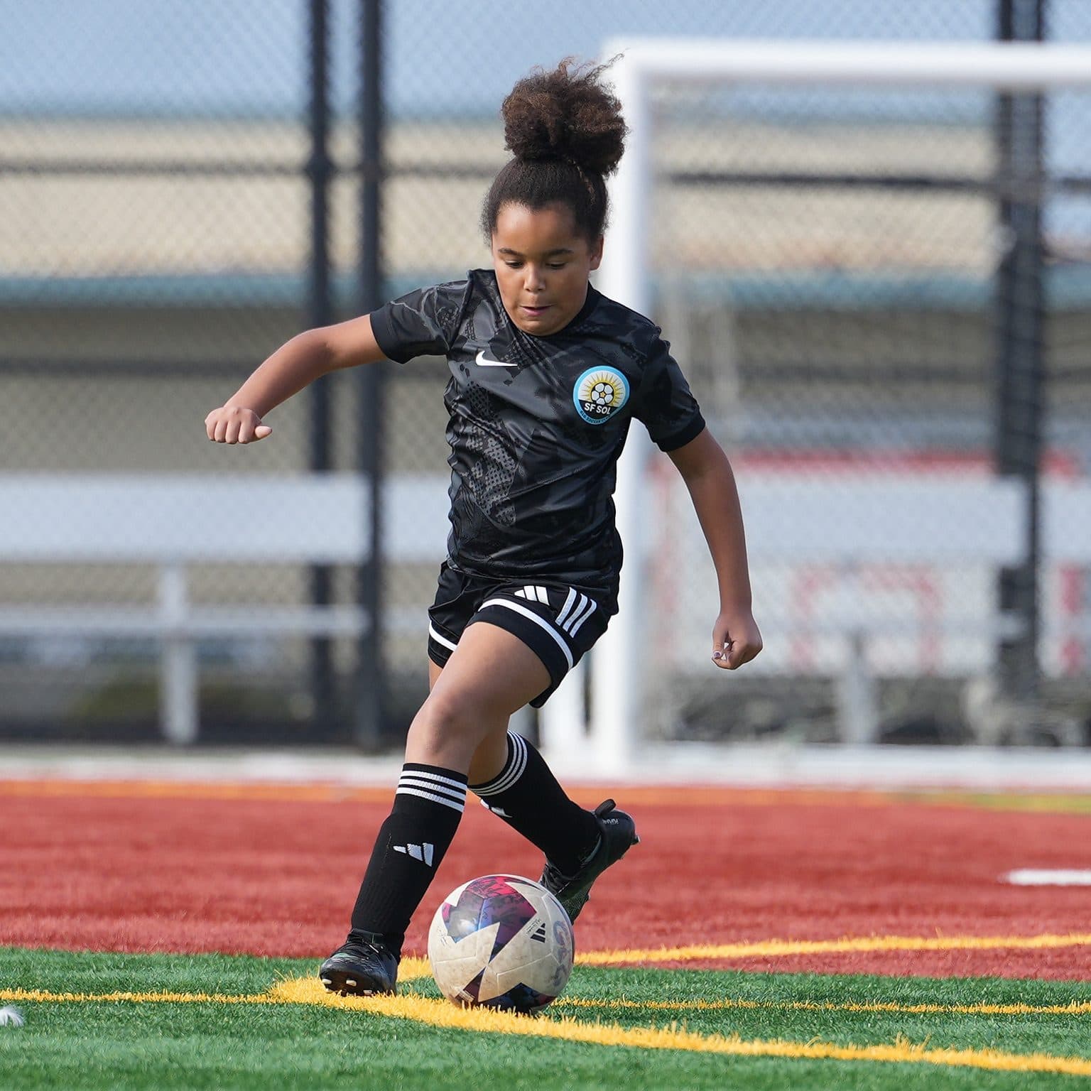 Vertical shot of a single GLG player in a black kit dribbling the ball down the turf, goal frame and fence behind her