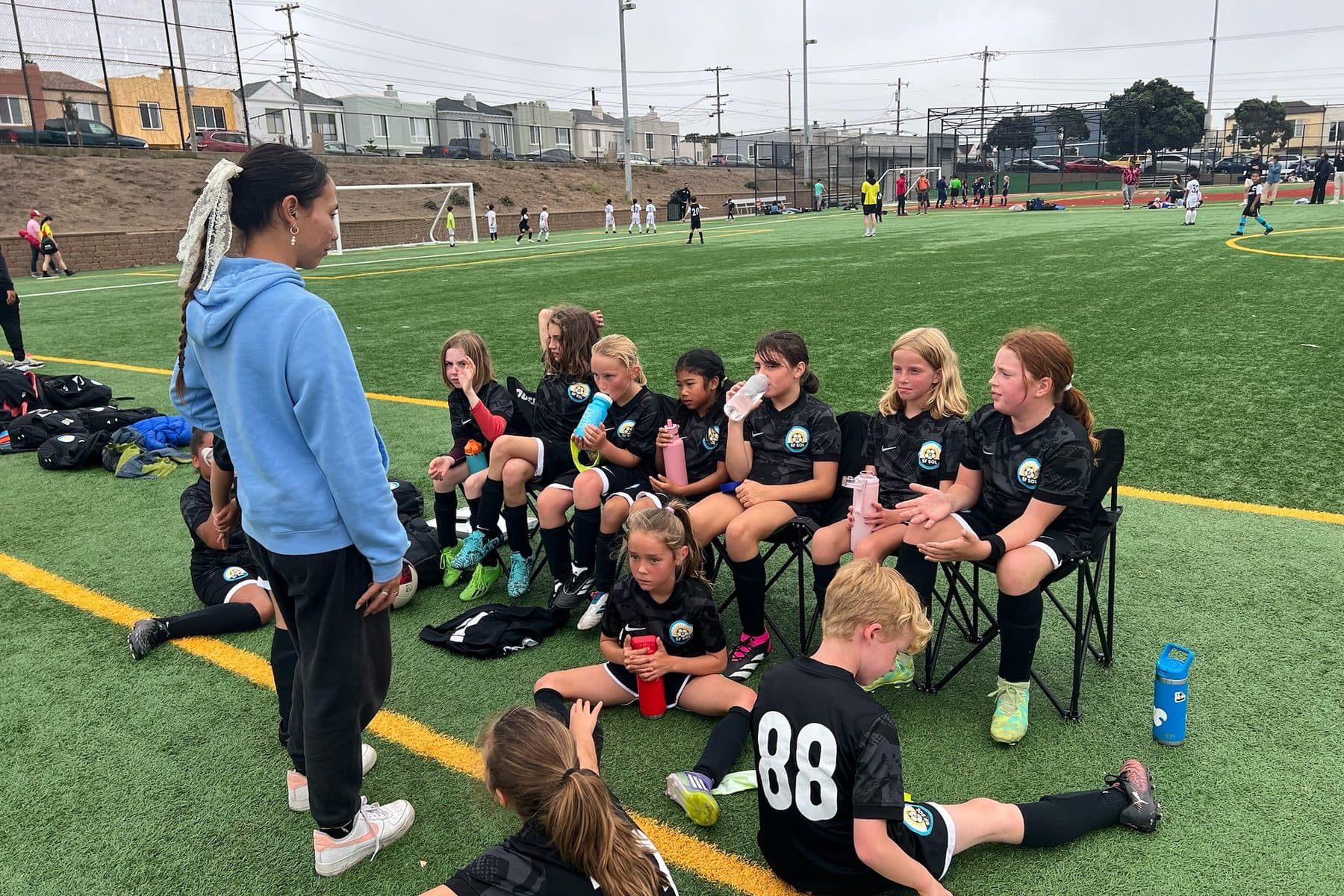 A coach in a light blue hoodie addresses players in black kits who sit in sideline chairs with water bottles, two kids stretching on the turf in front