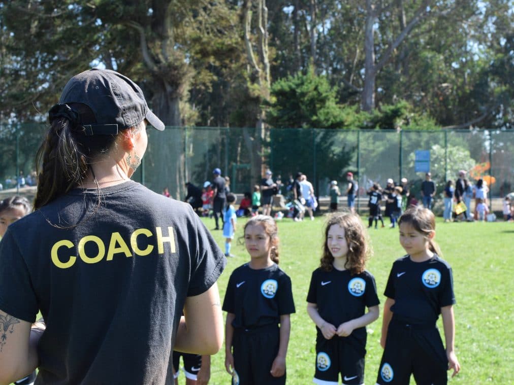Over-the-shoulder of a coach in a black 'COACH' tee talking to three girls in black GLG kits who listen in a line on a sunny grass field, busy sideline crowd blurred behind