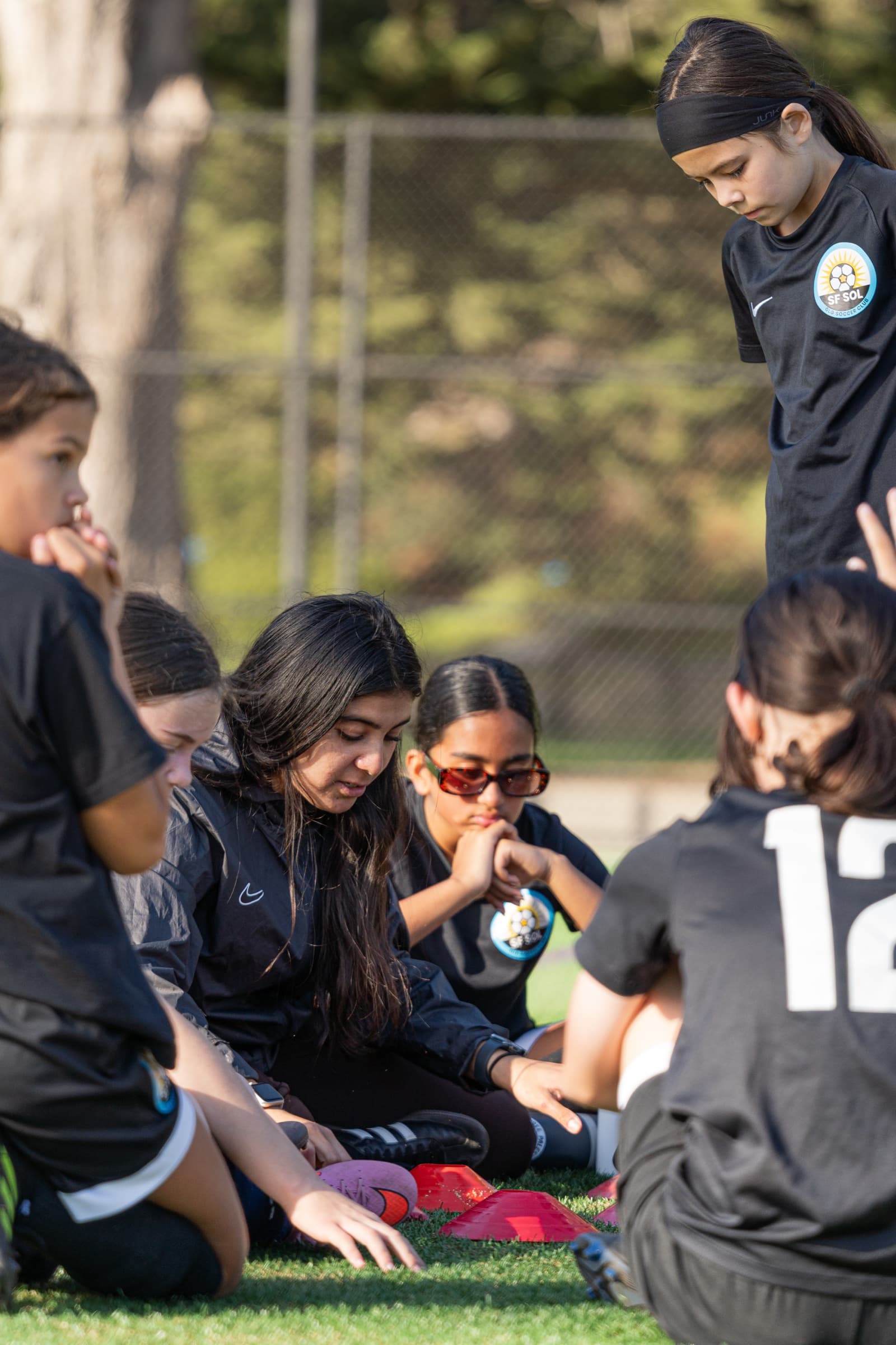 Players in black SF SOL kits sit in a huddle around red cones on the turf, one leaning in to point, while a teammate stands over them in golden light