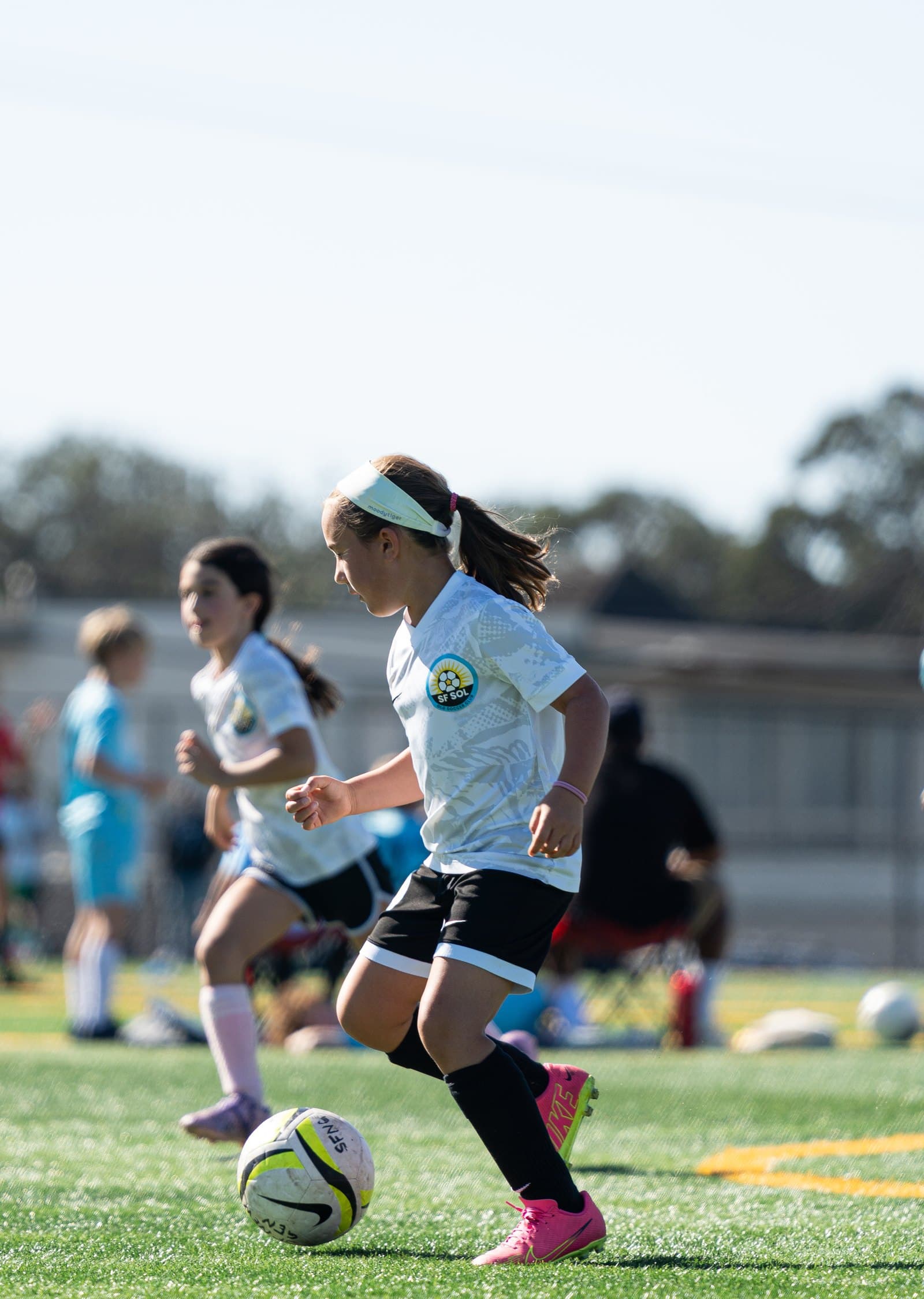 A player in a white kit and pink cleats dribbles across a sunny turf field, a teammate jogging behind