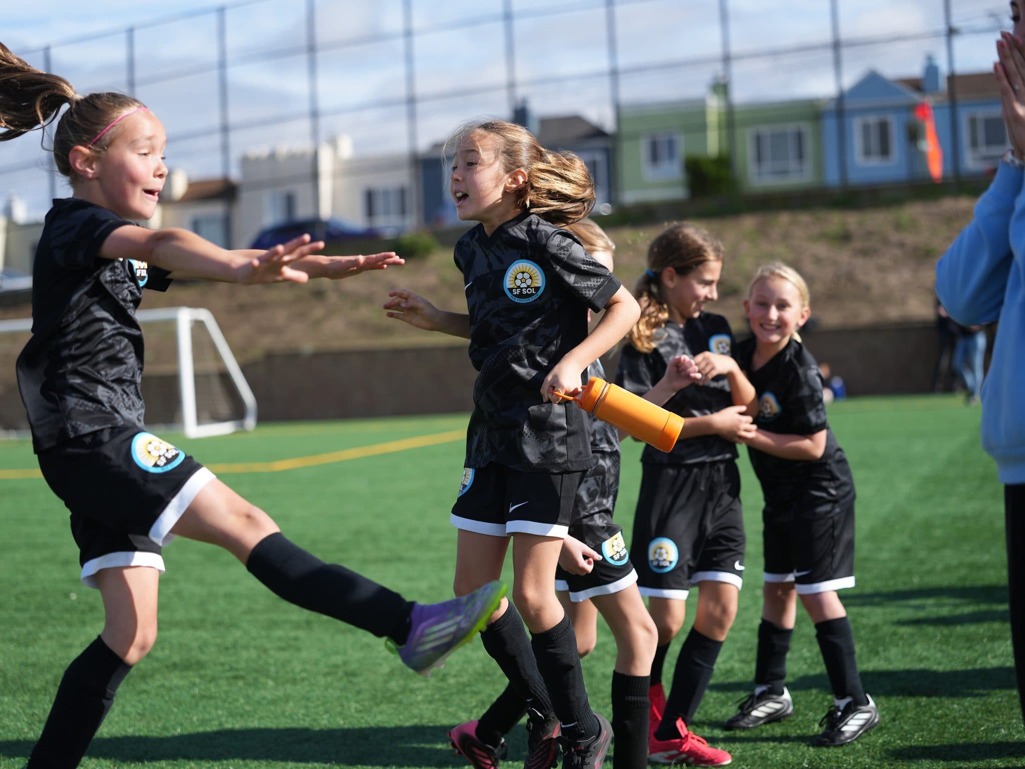 Pure joy: girls in black kits converge to celebrate on the sideline, one mid-leap with arms out, another beaming, a spectator clapping at frame right under blue sky with SF houses behind