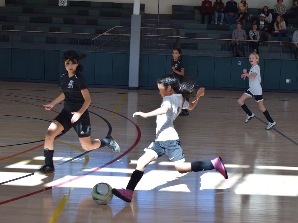 Indoor futsal action: a player in white loads up a big kick on the hardwood court as a defender in black sprints to close her down, spectators watching from the bleachers