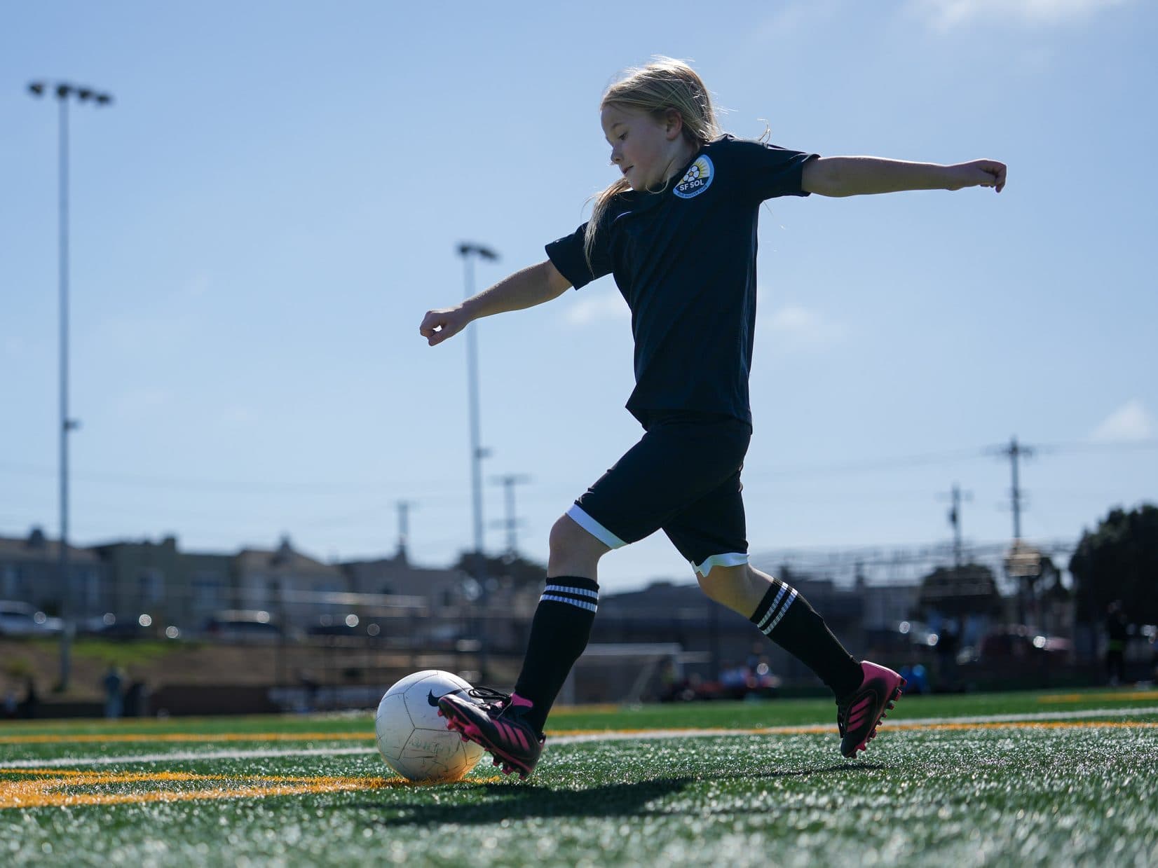 A lone player in a black kit plants her foot and swings through a shot on a sunny turf field, houses and light poles in the background