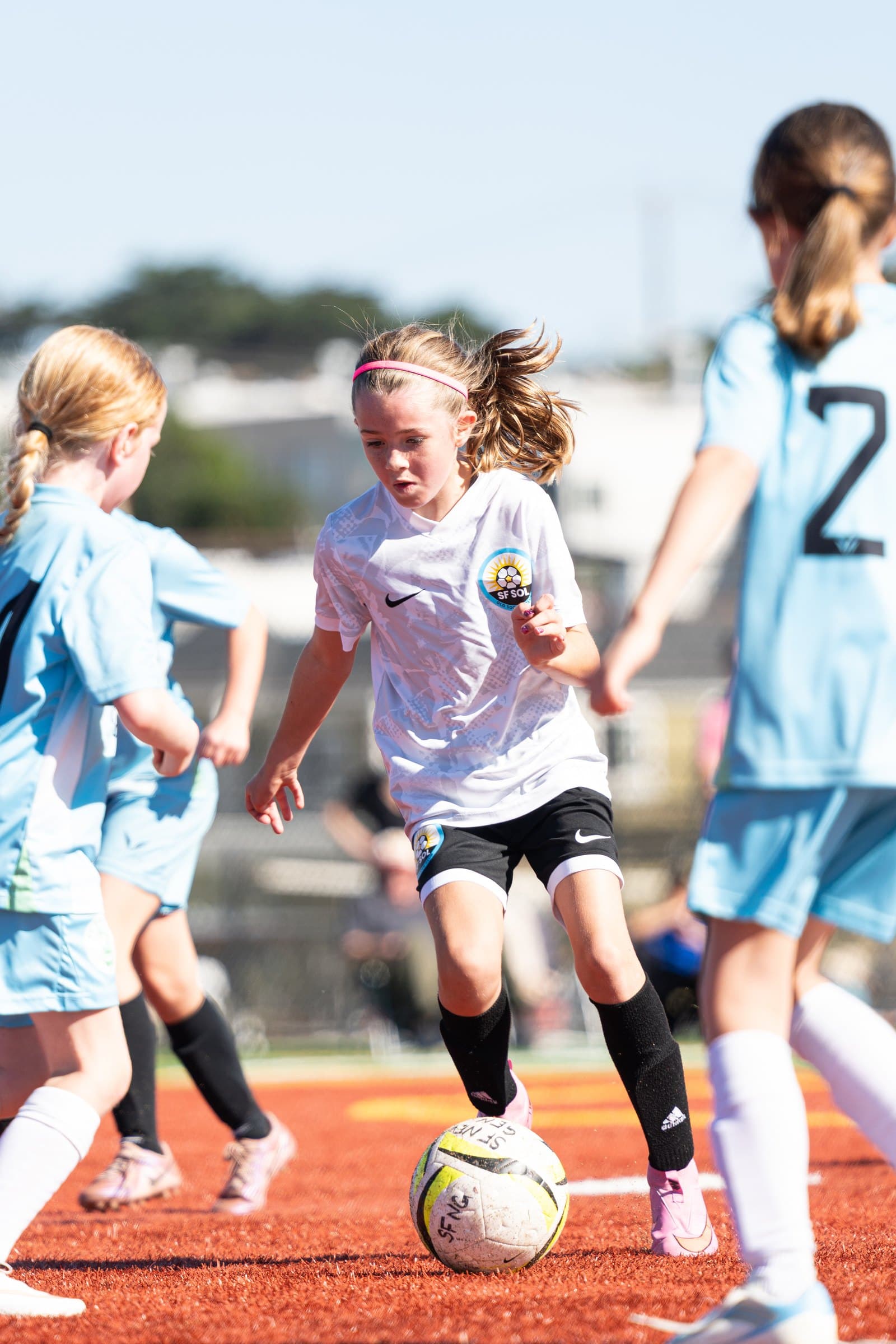 A player in the white away kit with a pink headband dribbles the ball on red turf, eyes down, as opponents in light blue close in on both sides