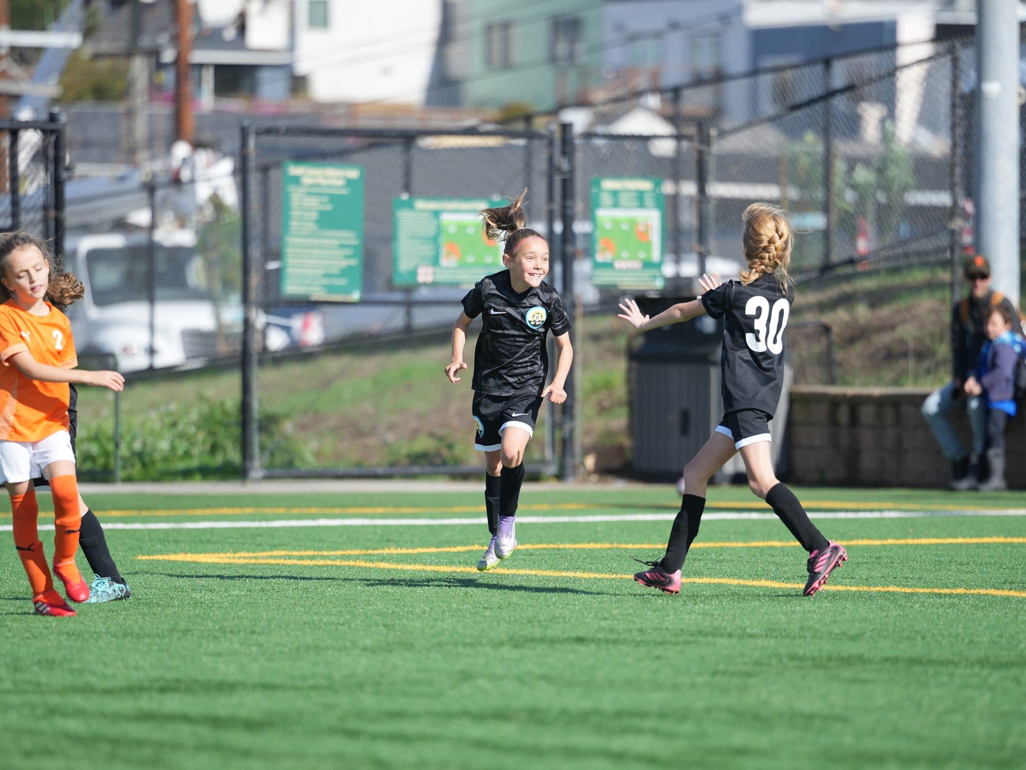 Two players in black kits (one wearing 30) run upfield during a game against an opponent in orange, the center girl grinning with ponytail flying