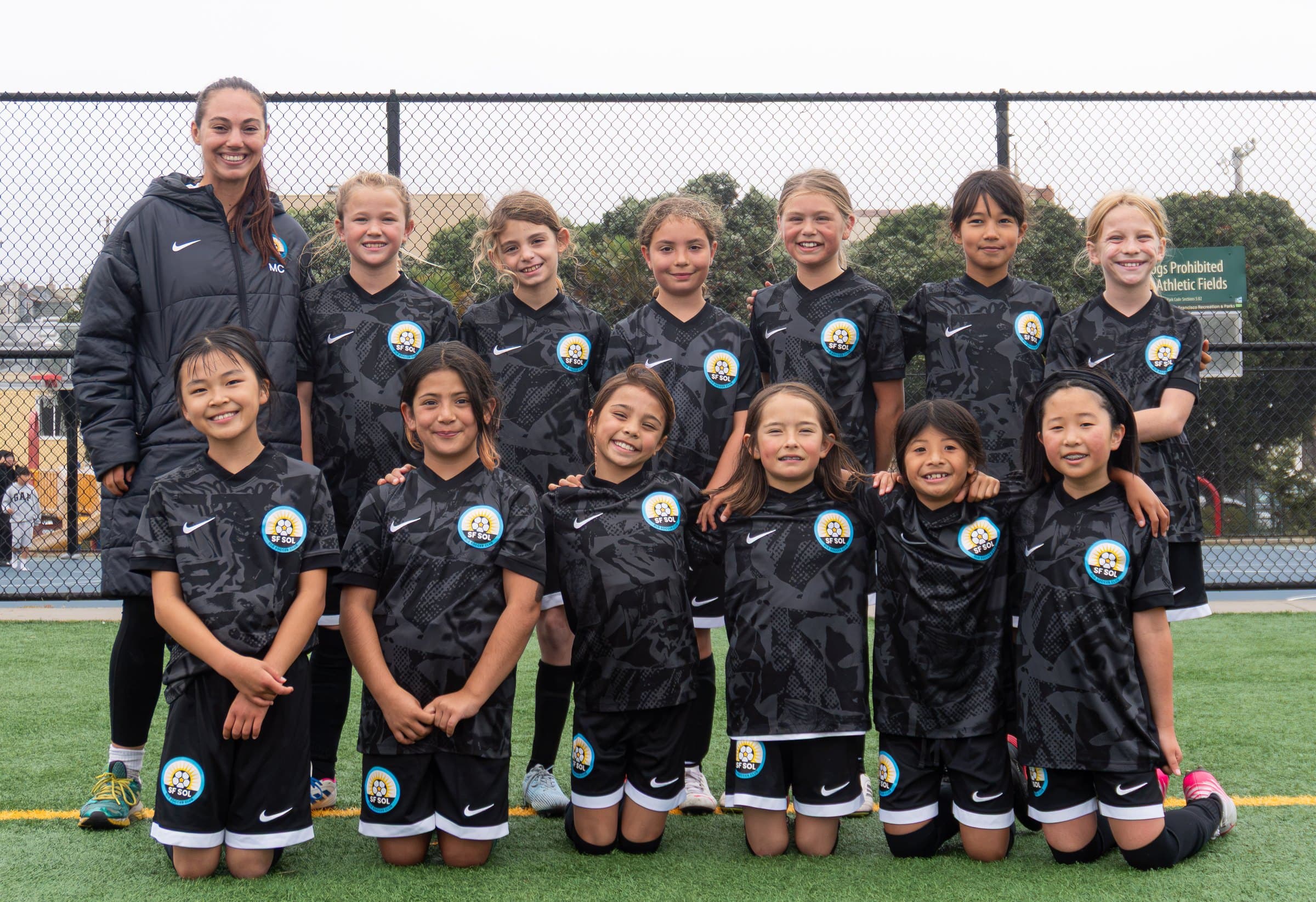 A team in black kits poses in two rows on the turf, front row kneeling, with their coach smiling at the left end of the back row