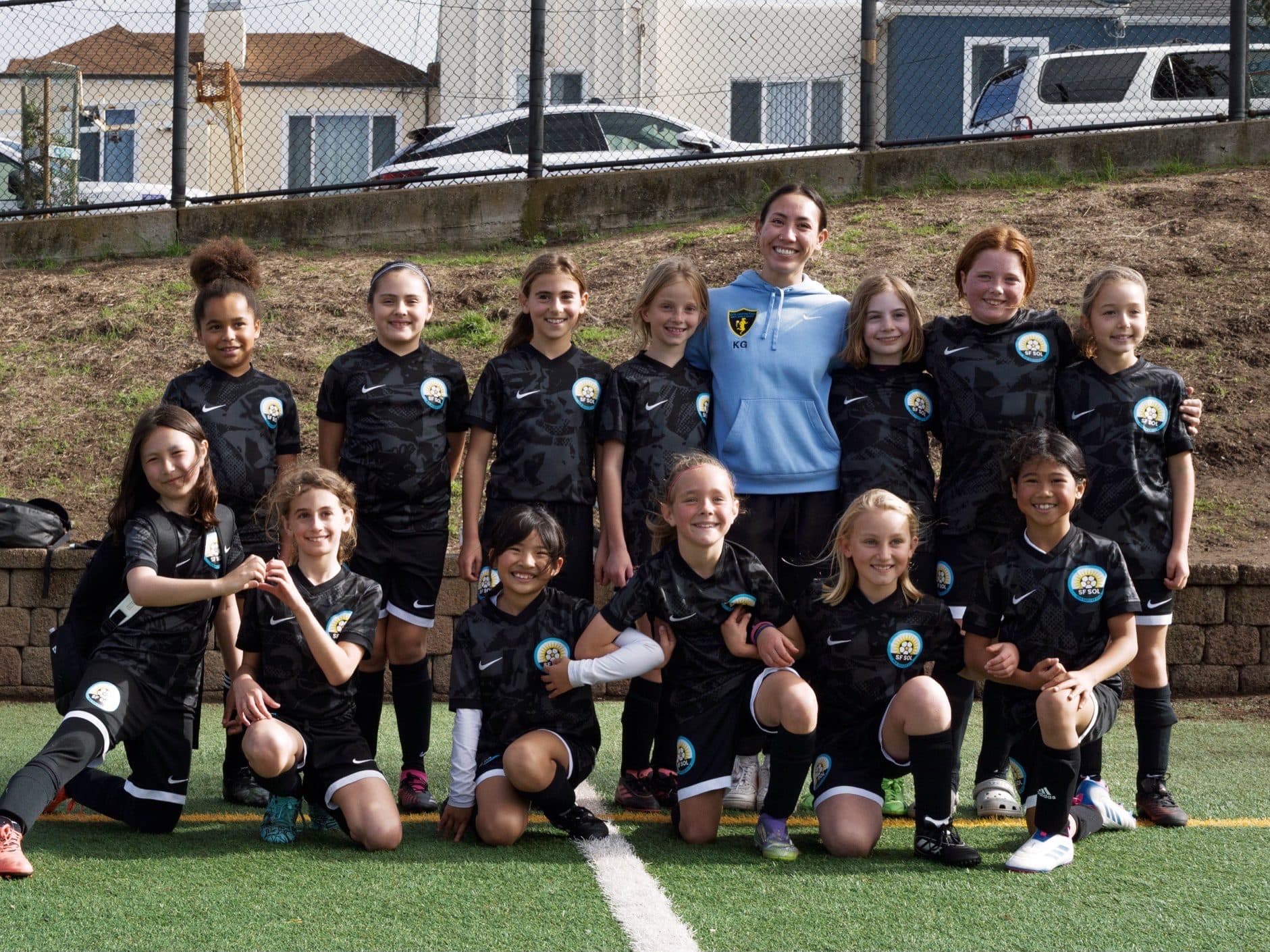 A full team in matching black kits poses in two rows on the turf sideline with their coach in a light blue hoodie standing at the center, everyone smiling
