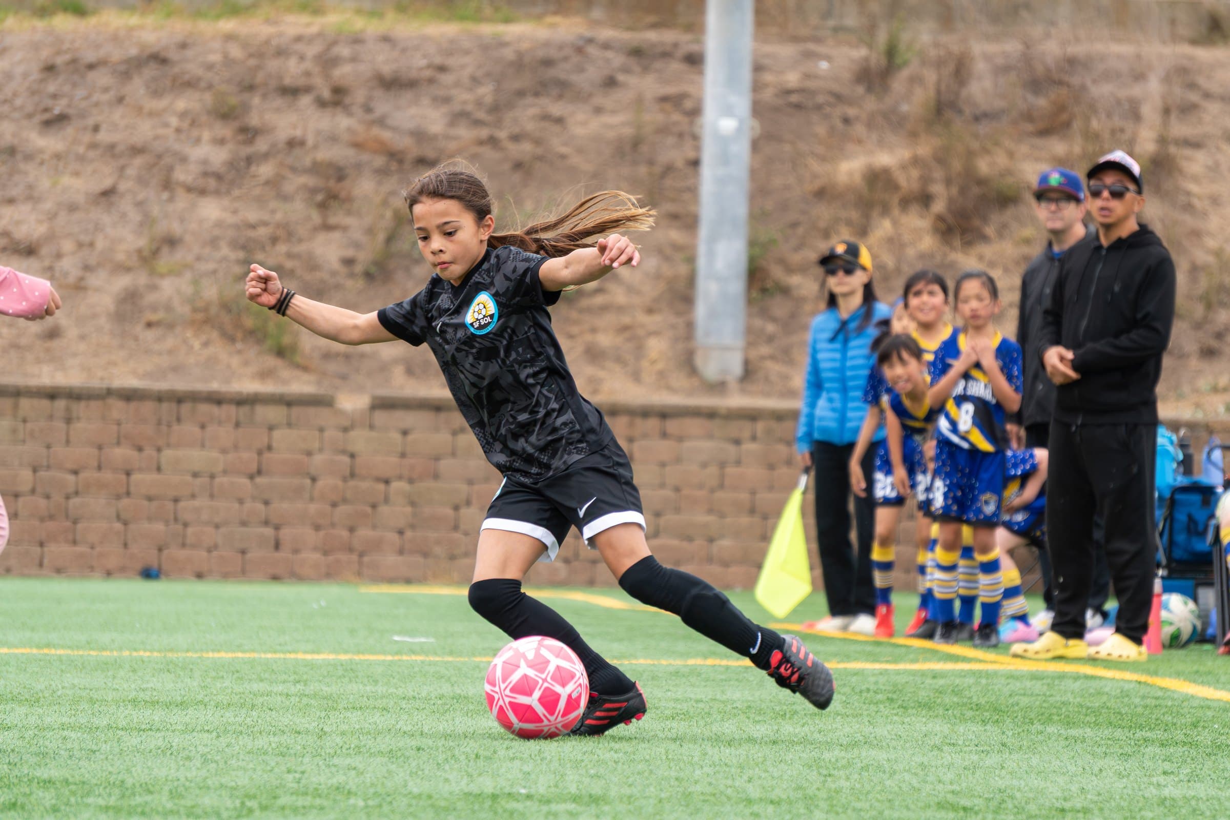 A player in the black club kit drives toward a pink ball on the turf, hair flying and arms out for balance, mid-stride