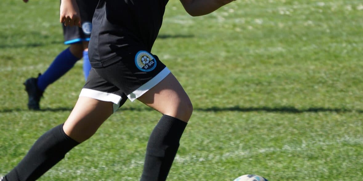 A determined player in a black kit with club crest drives onto the ball at full stride on a grass field, a defender in white and blue trailing behind her
