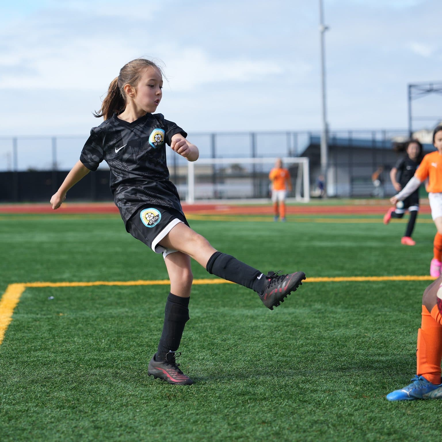 A GLG player in black is caught at full extension of a clearance or shot, ponytail flying, with orange-kitted opponents entering frame right