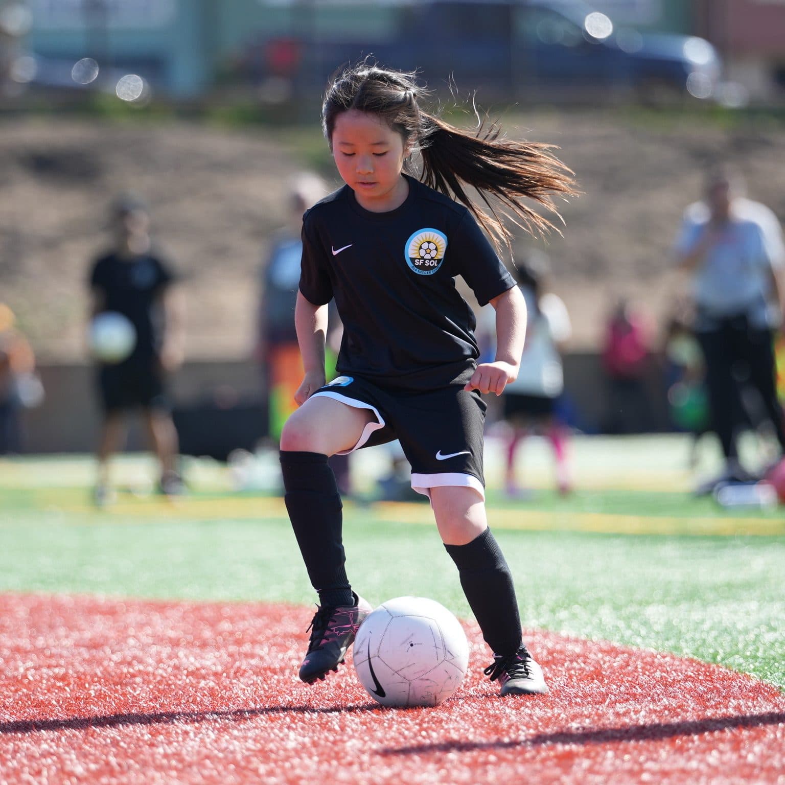 Sharp close action shot of a girl in the black club kit, hair flying, stepping on the ball to shield it from a rival in white/blue on sunlit red-and-green turf