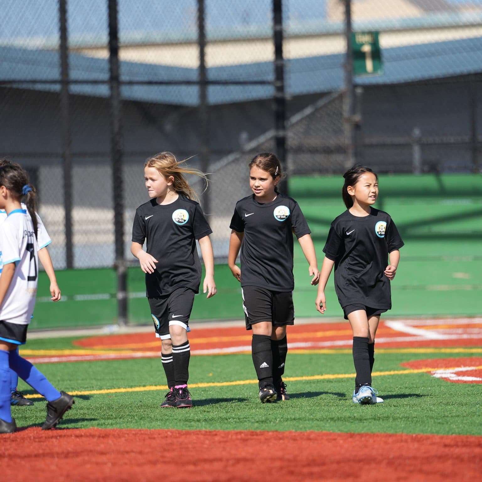 Three GLG players in black kits walk side by side across the turf during a break in play, with white-kit opponents at the left