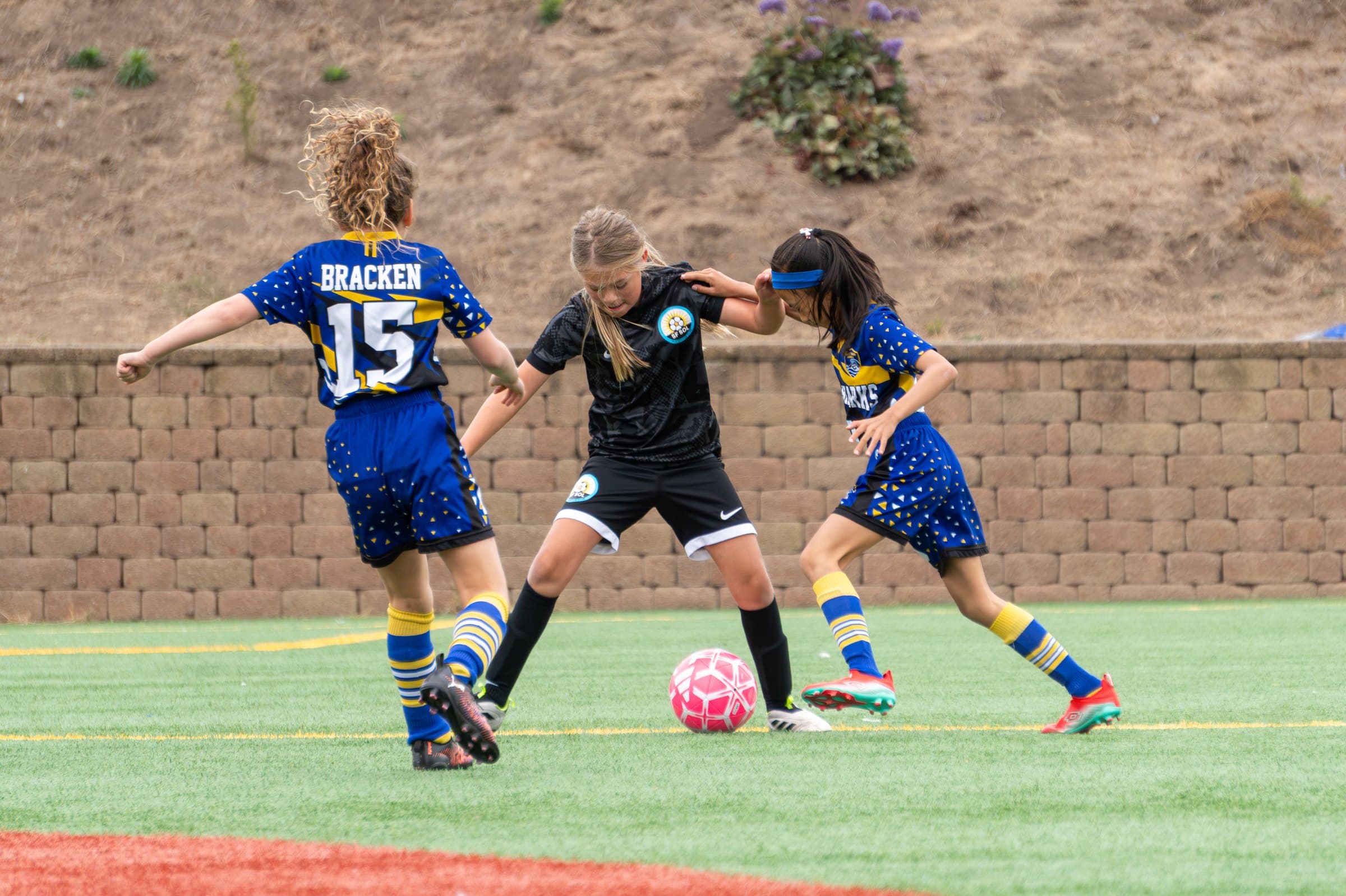 A player in the black club kit shields a pink ball between two defenders, arms out and eyes on the ball