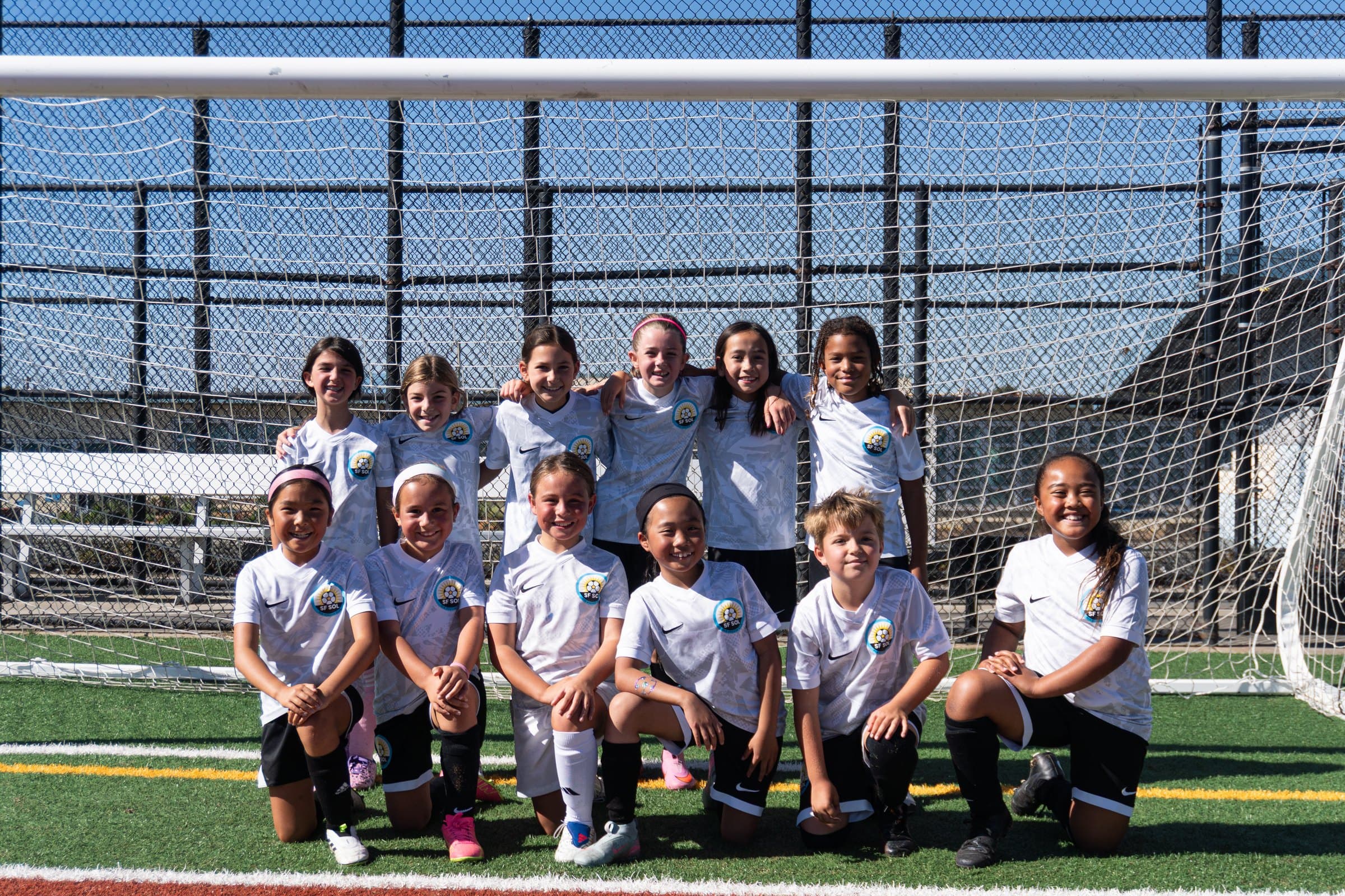 A team in white kits poses in front of the goal, back row arm in arm and front row kneeling, all grinning