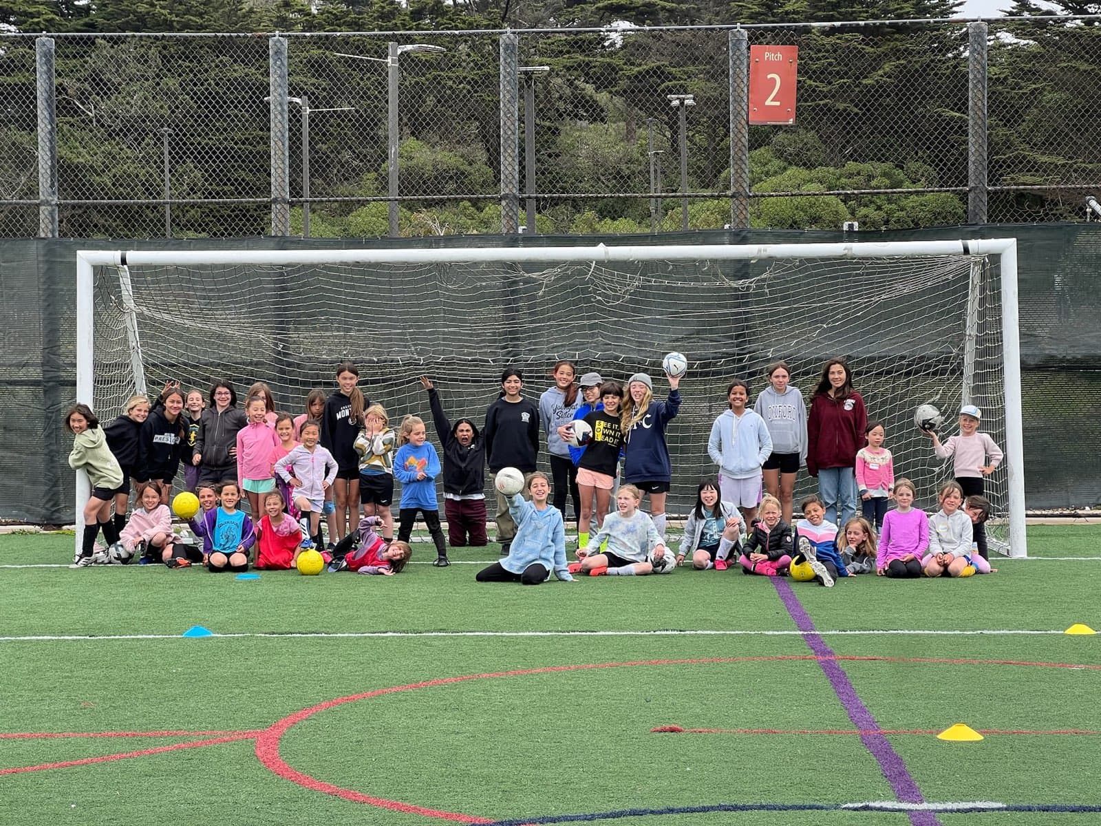 About forty campers in hoodies and T-shirts crowd in front of a full-size goal on Beach Chalet's Pitch 2, some kneeling with balls held high, others waving and mugging for the camera