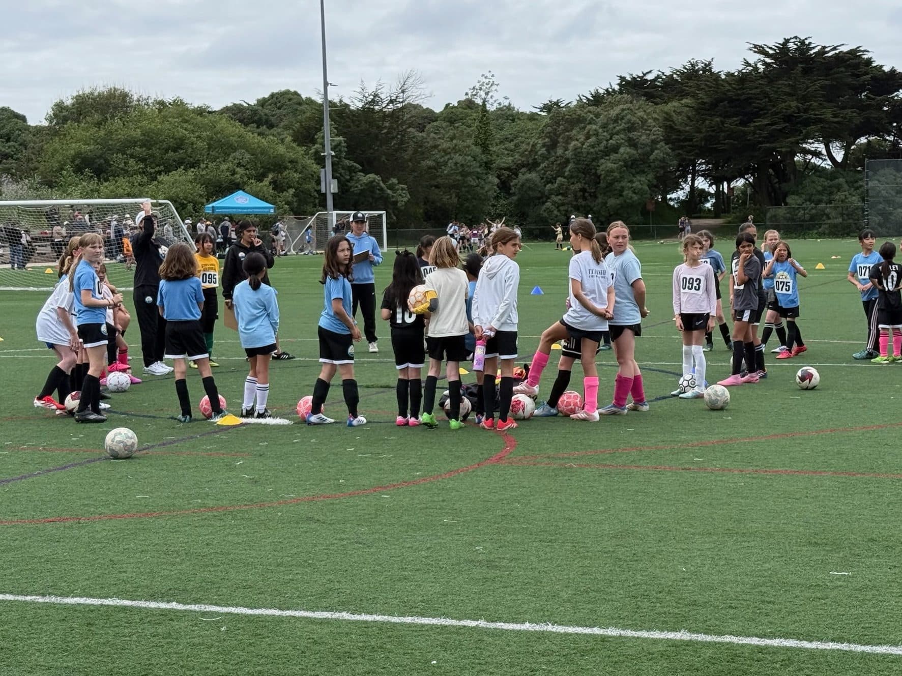 A big crowd of young players in light blue tees and white shirts with pinned paper numbers stands among cones and pink balls on an overcast turf field