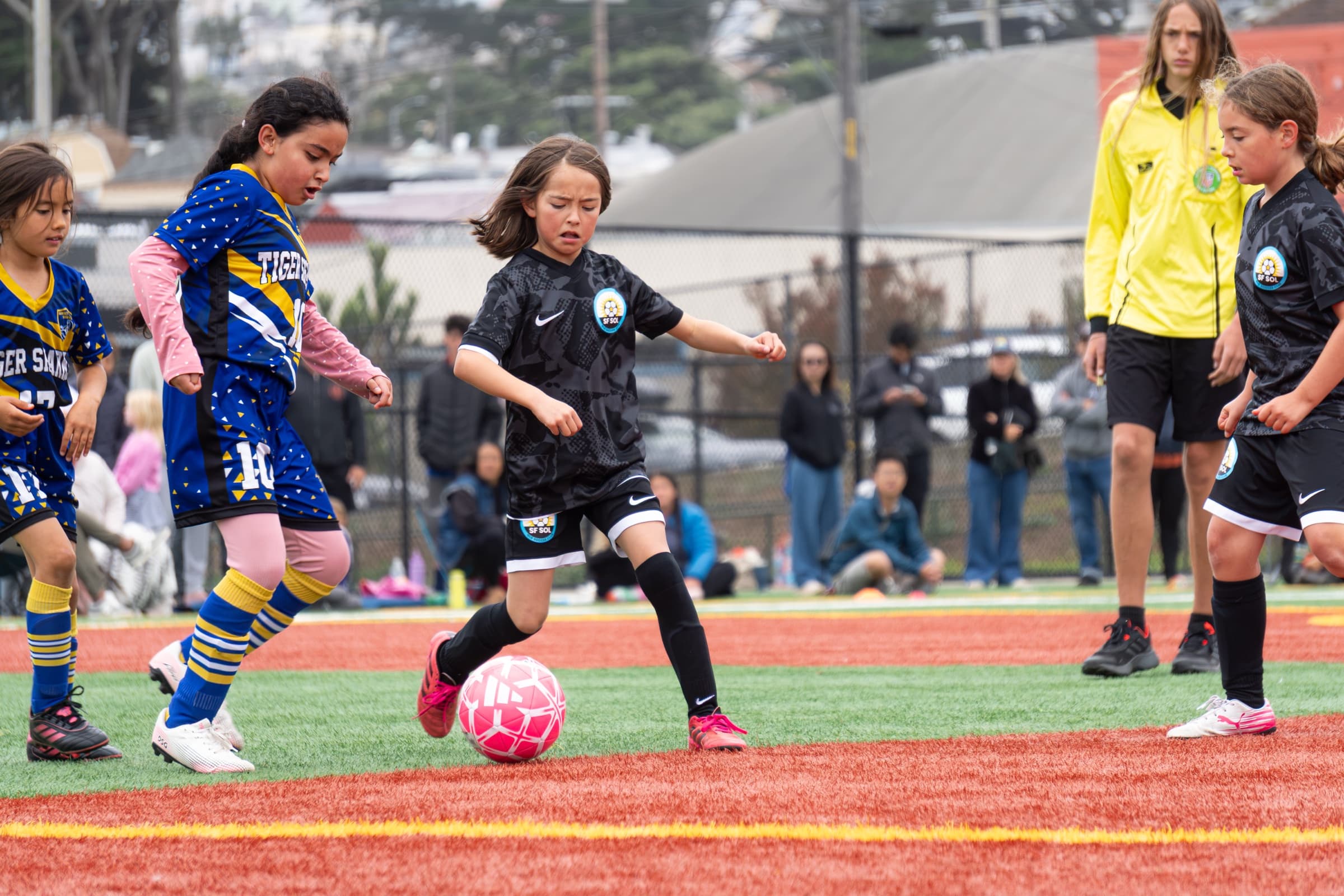 A player in the black SF SOL kit dribbles a pink ball upfield, eyes ahead, as opponents in blue and gold close in and a referee in yellow watches