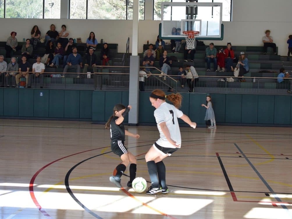 A GLG player in black challenges a white-shirted #7 for the ball on a hardwood gym floor, with families watching from the bleachers under basketball hoops