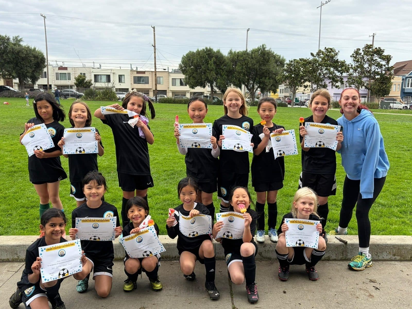 Two rows of players in black kits proudly hold up certificates while a smiling coach in a blue hoodie stands at the right, city houses behind the park