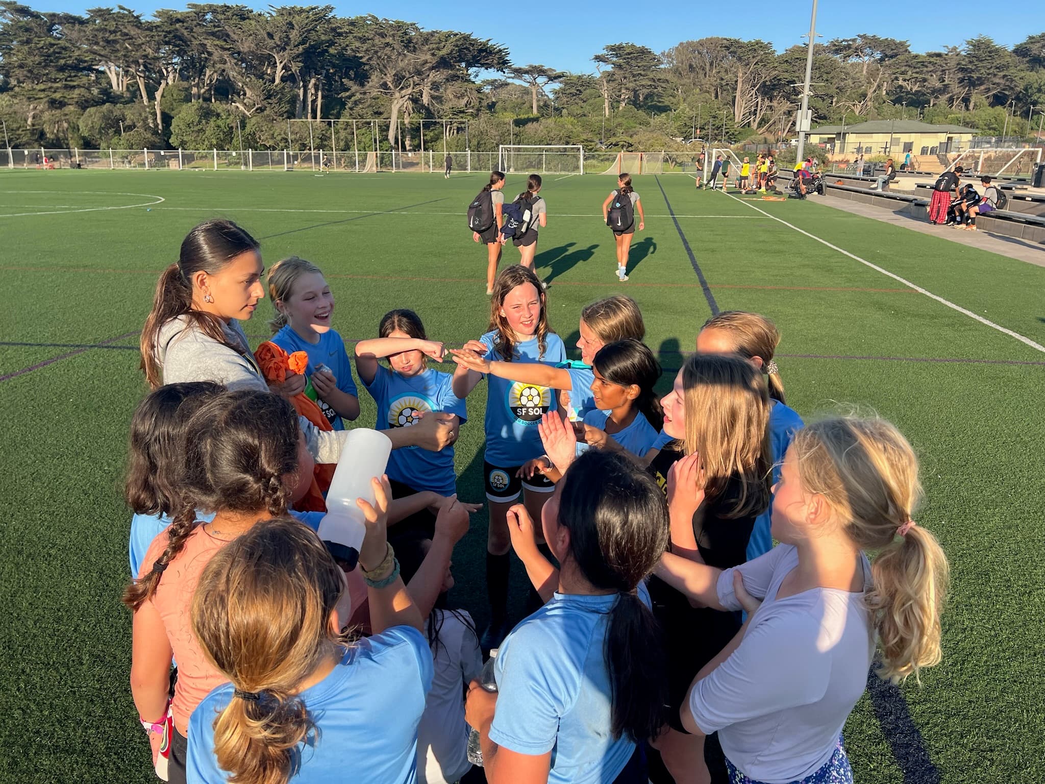 A rec team in light blue shirts crowds into a hands-in huddle with their coach in golden-hour light, cypress trees behind the field