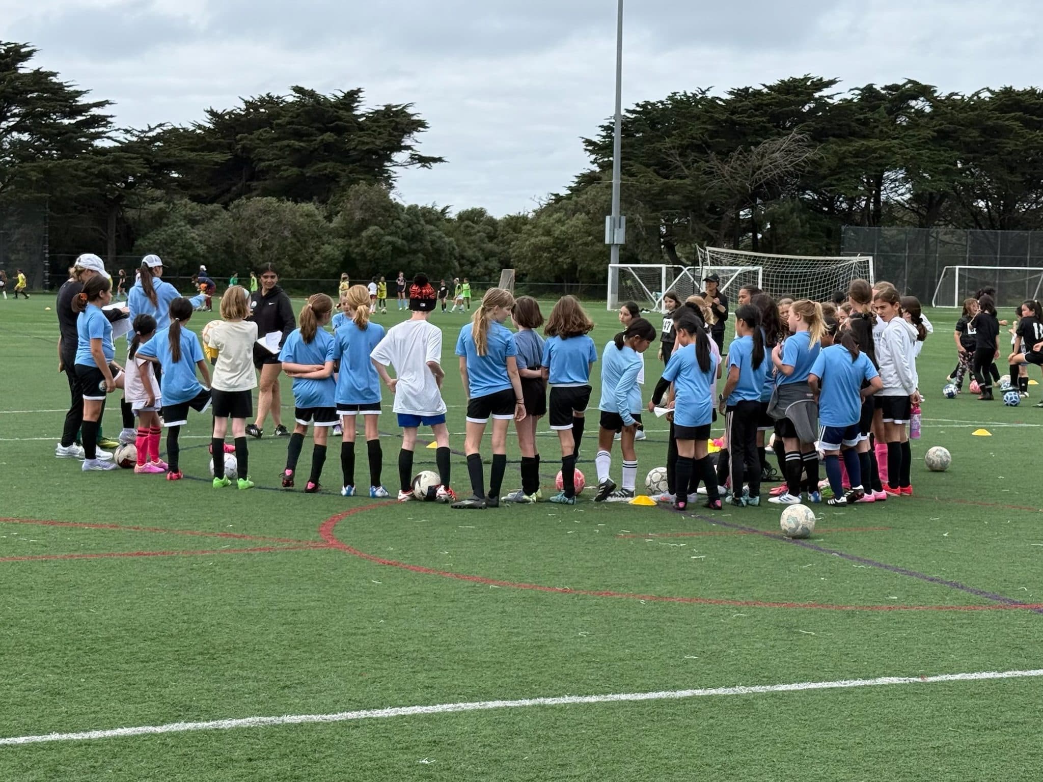 Wide shot of a big rec practice: dozens of girls in light blue shirts form a circle around coaches on a turf field ringed with cones and balls