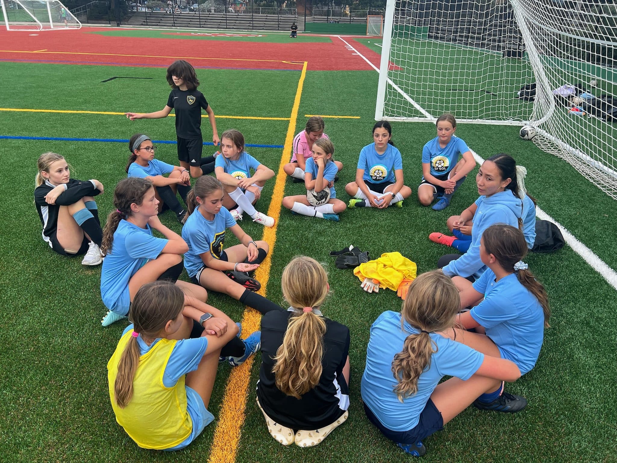End-of-practice circle: a dozen players in light-blue jerseys (a few in yellow pinnies) sit on the turf near the goal while a coach in black stands and talks, evening light going flat