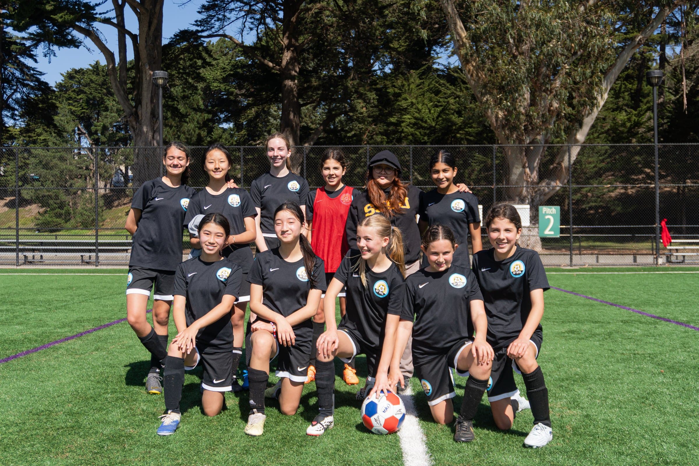 A team in black kits poses in two rows on a sunny turf field, front row kneeling around a ball, tall eucalyptus trees behind