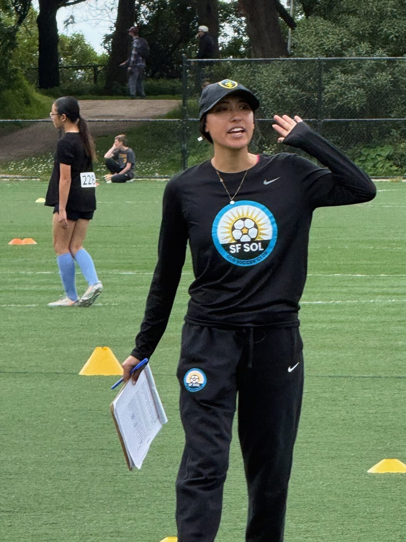 A smiling coach in black SF SOL branded top and bucket hat walks the turf with clipboard in hand, gesturing instructions, yellow cones and players in the background