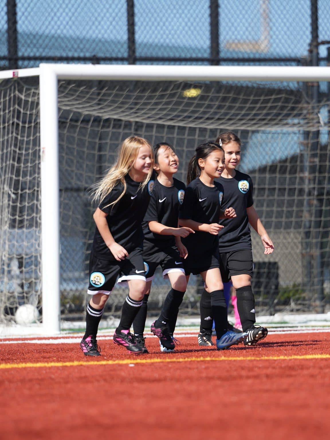Four smiling teammates in black kits stride away from the goal together in celebration, with opponents in white and blue in the background on a sunny turf field