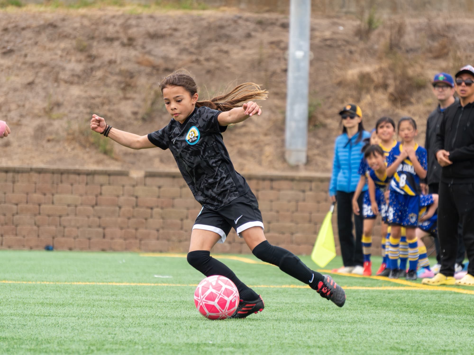 A player in a black SF SOL kit winds up to strike a pink ball, hair flying behind her, as the opposing bench watches from the sideline